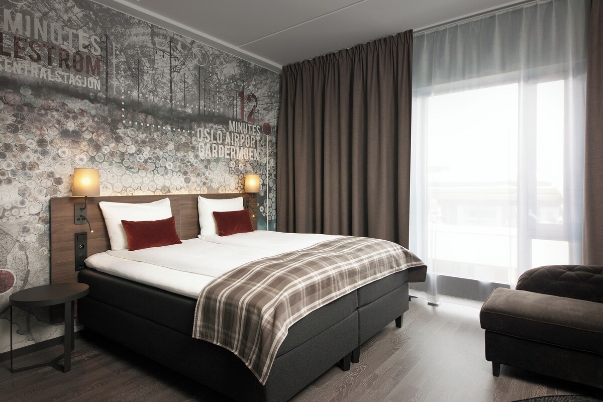 Habitación de hotel moderna con una cama grande, cabecero con reloj, una mesita y vistas a la ciudad a través de cortinas translúcidas.