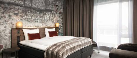 Una habitación de hotel moderna con una cama grande, una mesita auxiliar pequeña, un sofá y un ventanal con cortinas.
