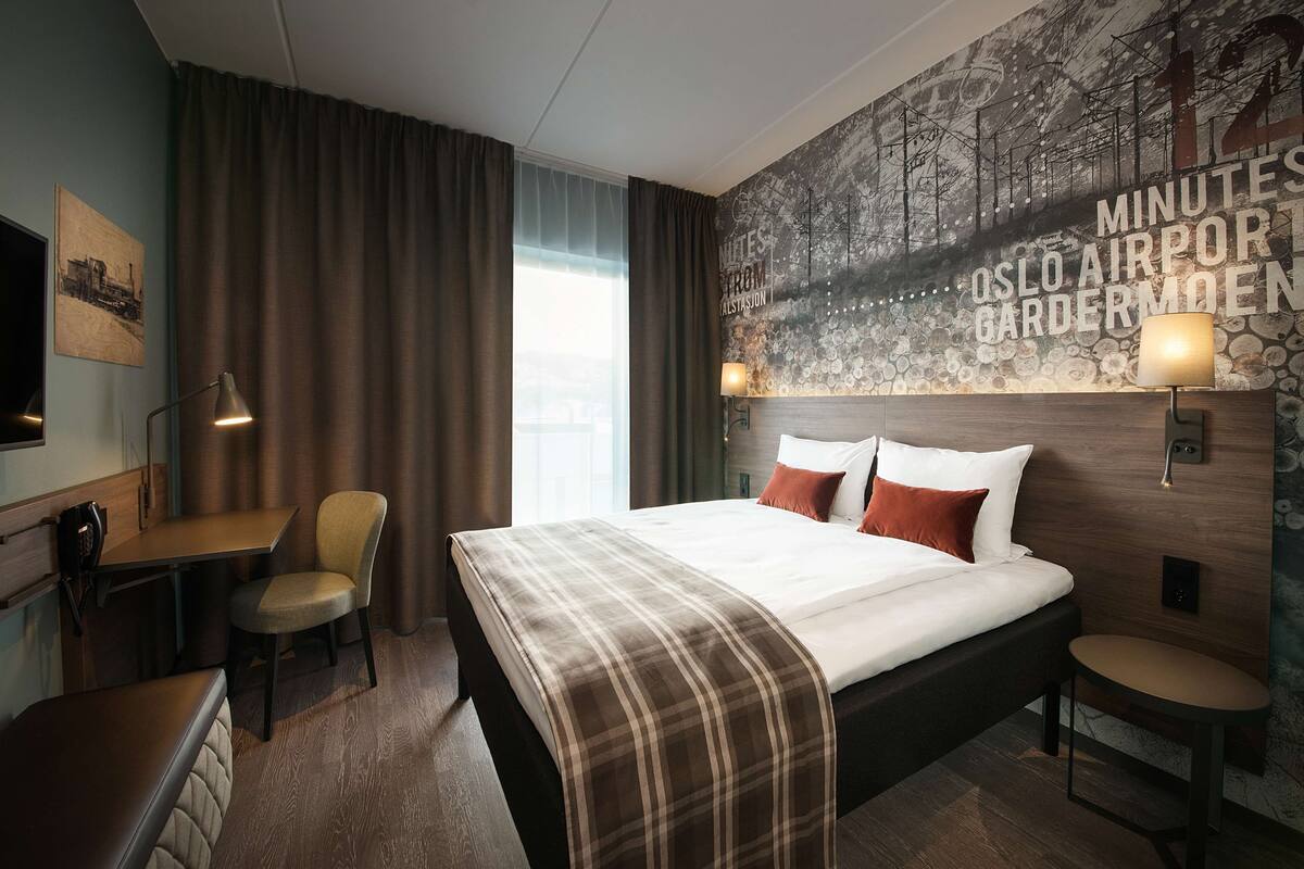 A hotel room with a bed, a desk, a chair, a small table, and a wall with a cityscape mural.