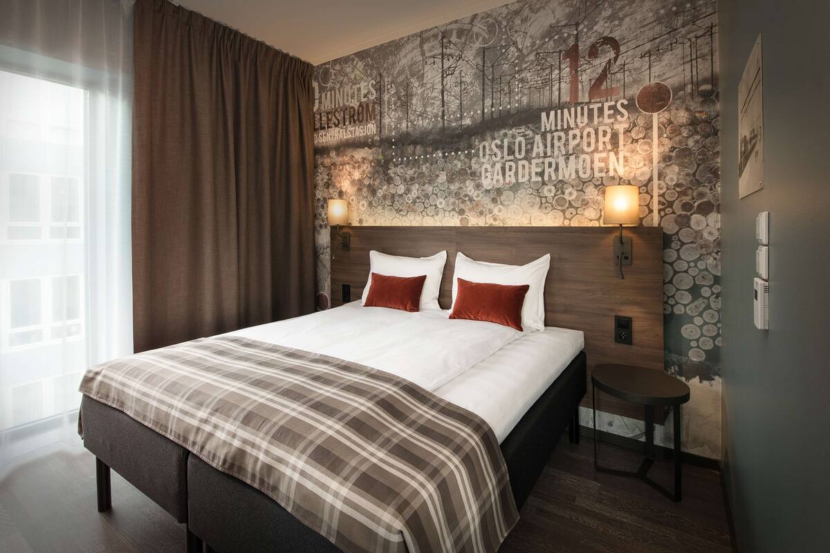 A modern hotel room with a large bed, two bedside tables, a headboard with a clock, and a mural on the wall.