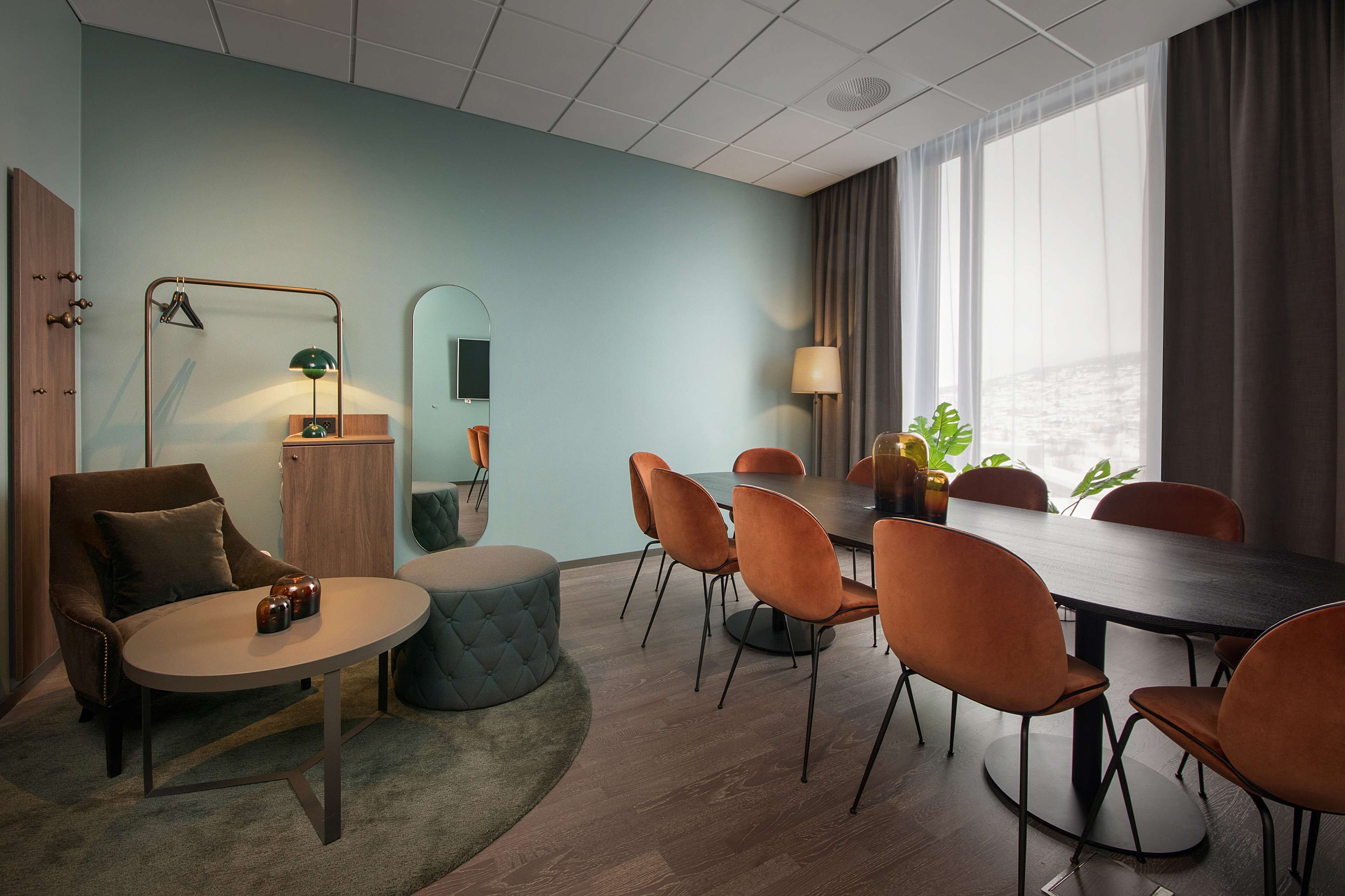A modern conference room with a round table, orange chairs, a tufted ottoman, a mirror, and a view of the outdoors through large windows.