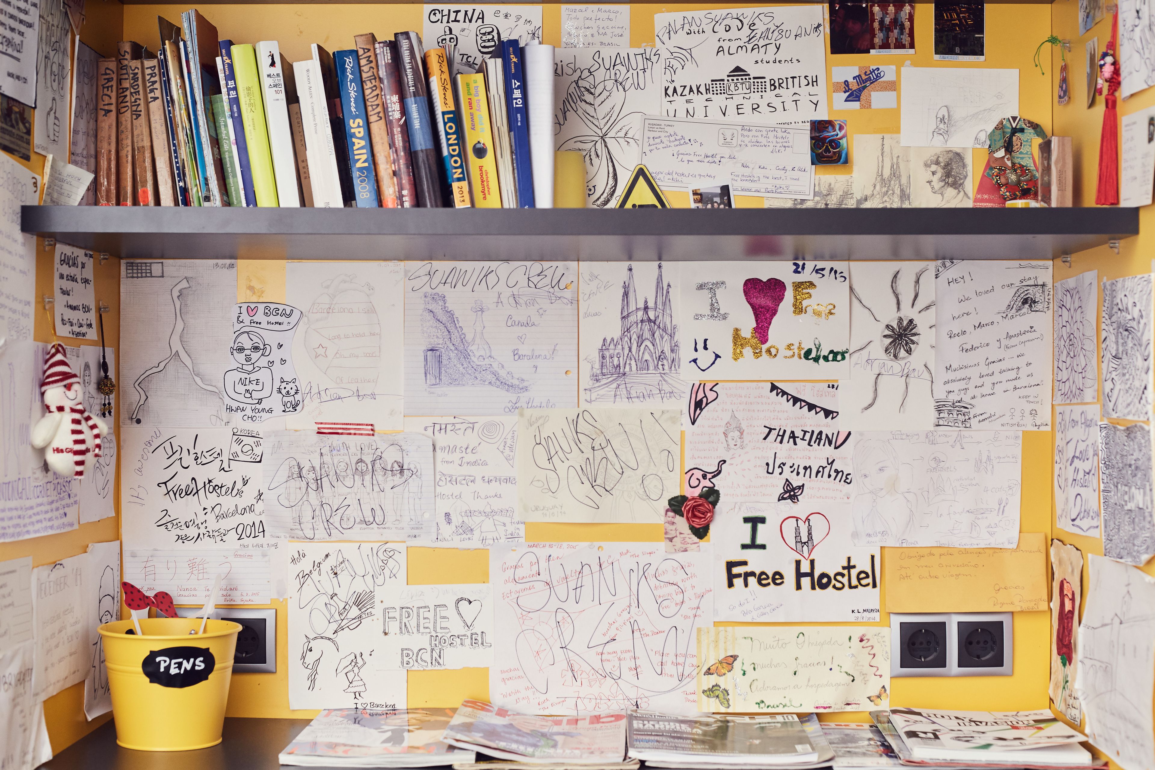 Een rommelige kamer met een boekenplank, verschillende papieren en een gele emmer met de tekst 'PENS'.