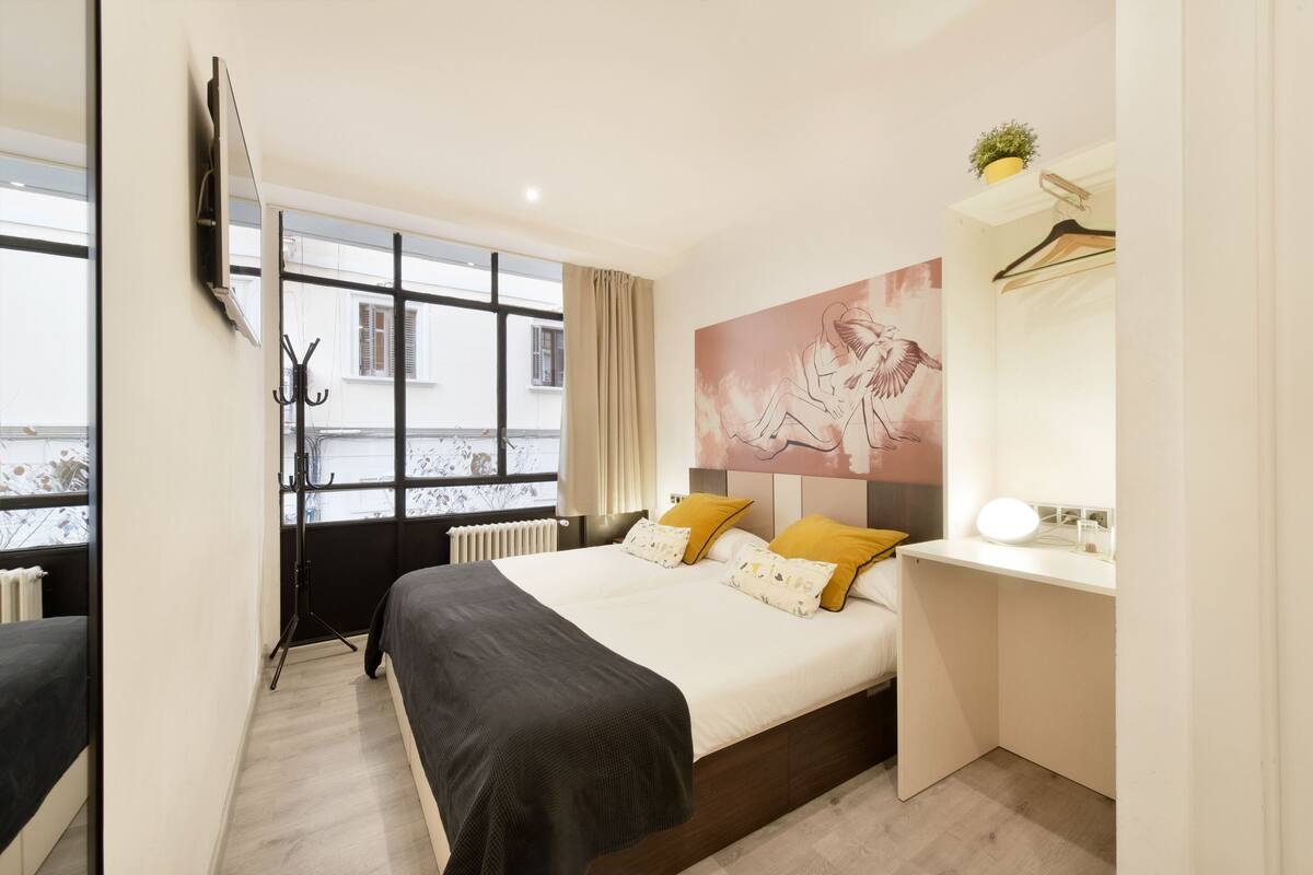 Ein modernes Hotelzimmer mit einem großen Bett, einem Schreibtisch, einem an der Wand befestigten Fernseher und einem Fenster mit Blick auf Gebäude.