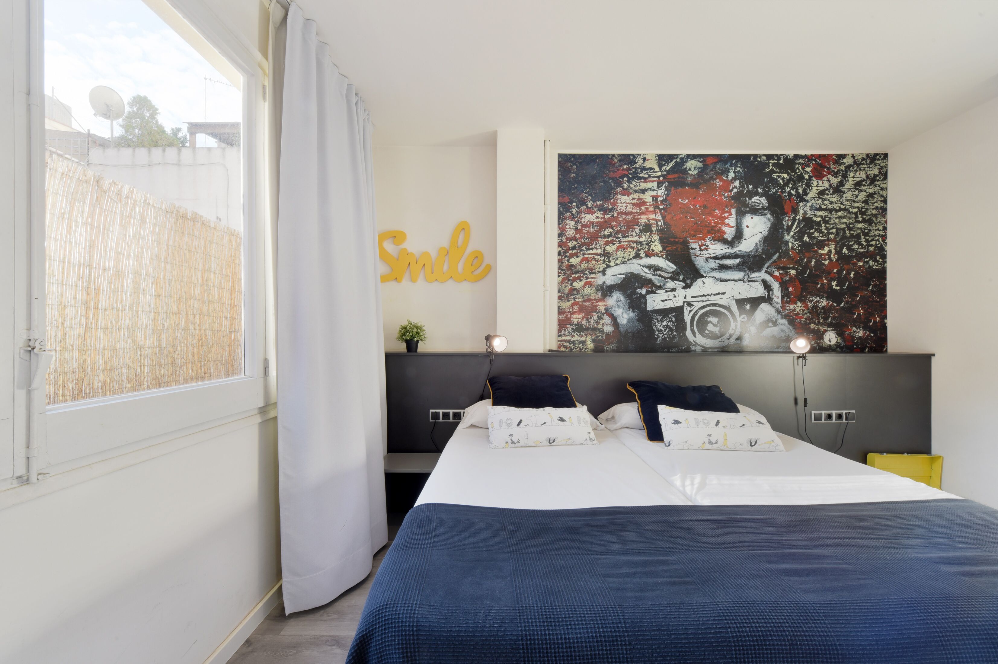 A bedroom with a bed, a wall-mounted TV, a large painting, and a window with a curtain.