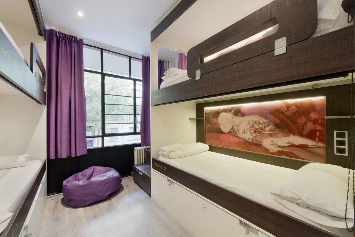 Ein modernes Hotelzimmer mit Etagenbett, Sitzsack, großem Fenster mit violetten Vorhängen und einem an der Wand befestigten Fernseher.