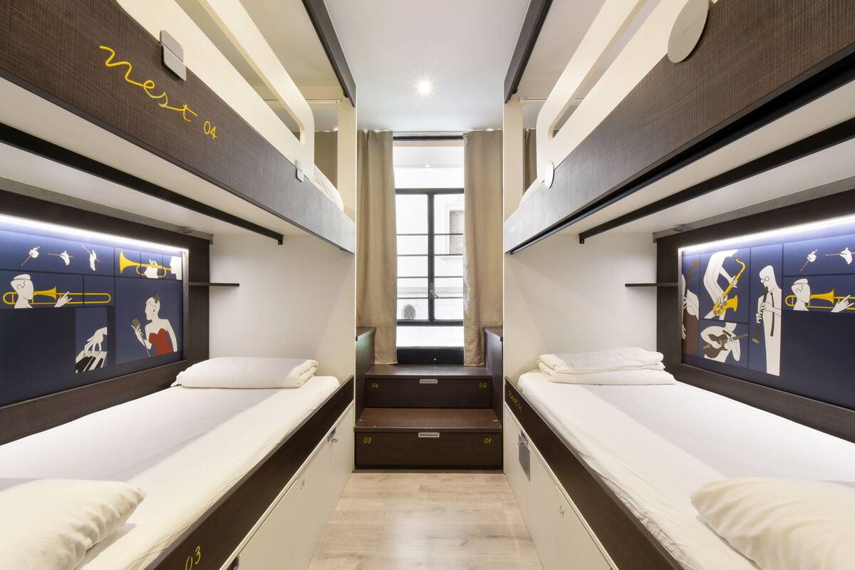Ein schmales Zimmer mit zwei Betten, beide mit Kopfteilen, die musikalische Instrumente zeigen. In der Mitte ist ein Fenster mit Vorhängen zu sehen.