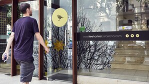 A man walking towards a building with a glass entrance, a yellow circular sign with a bird, and a website address on the glass. - Free Hostels Barcelona (Barcelona)