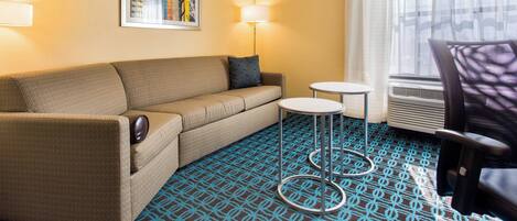 A hotel room with a beige sofa, a small round table, a chair, a lamp, and a patterned carpet.