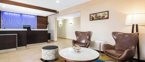 A lobby with a reception desk, two leather armchairs, a round table with a bowl, a small round table, and a lamp.