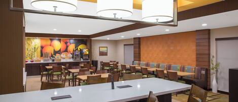 A hotel dining area with multiple tables and chairs, a counter with a fruit display, and a seating area with a patterned wall.