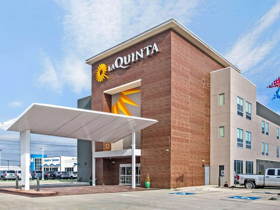 Um hotel La Quinta com design moderno, apresentando um logotipo solar em destaque, área de estacionamento coberta e céu claro.