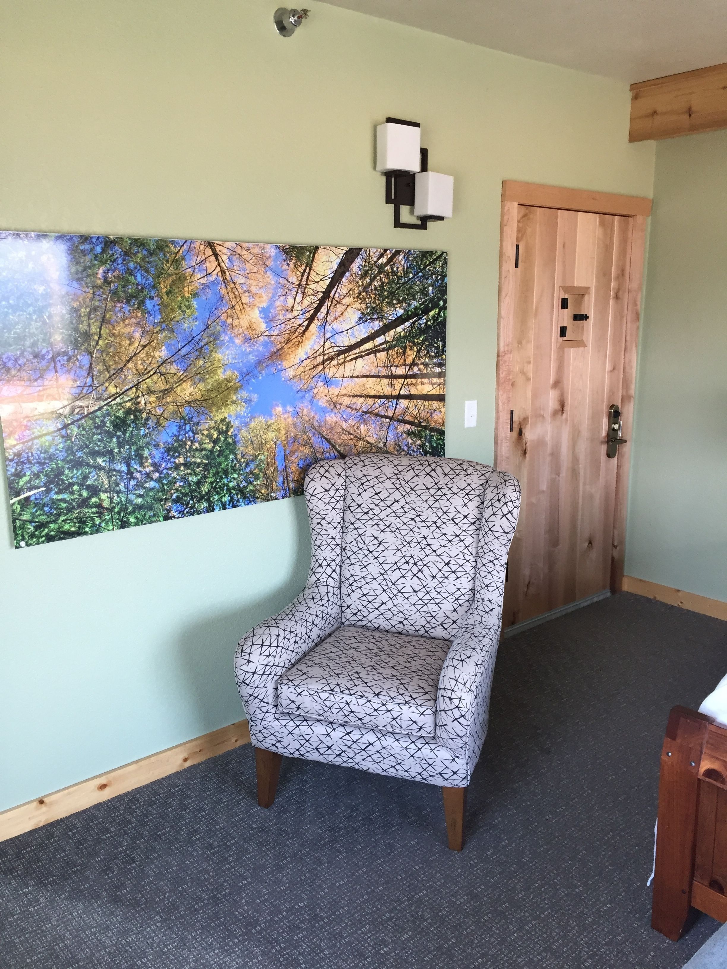 A room with a patterned armchair, a large nature-themed wall hanging, a wooden door, and a small wooden table.