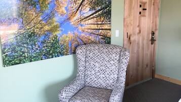 A room with a patterned armchair, a large nature-themed wall hanging, a wooden door, and a small wooden table.