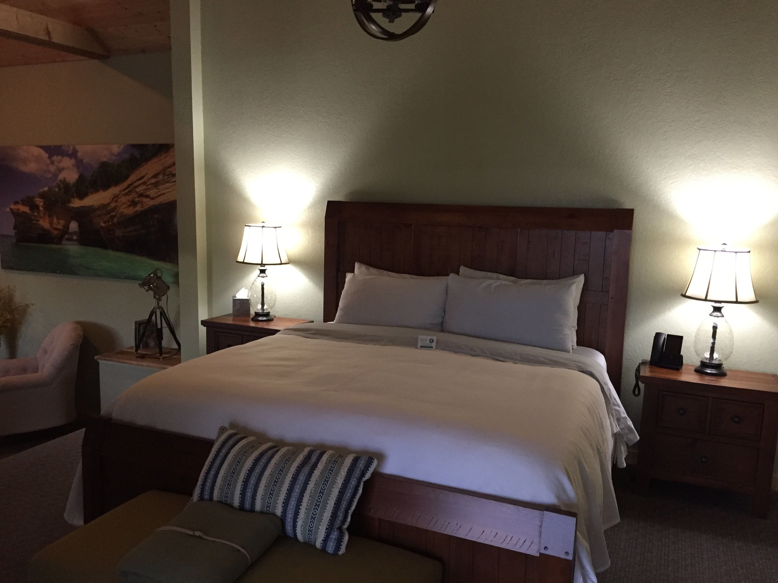 A neatly made bed with a wooden headboard, two bedside lamps, and a small bedside table with a lamp and a camera on it.