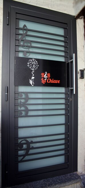 A black metal door with a frosted glass panel, featuring a treble clef and musical notes design, and a sign reading "B&B Chiave".
