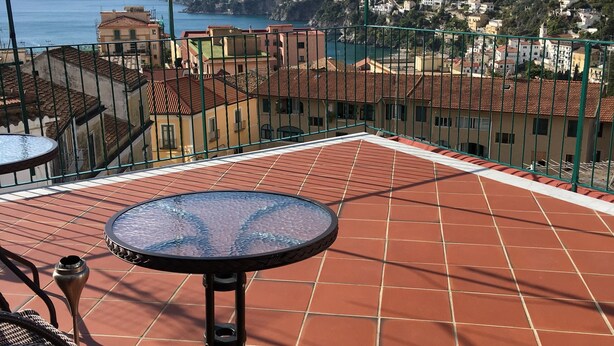 A terrace with a round table and wicker chairs, overlooking a coastal town and mountains.