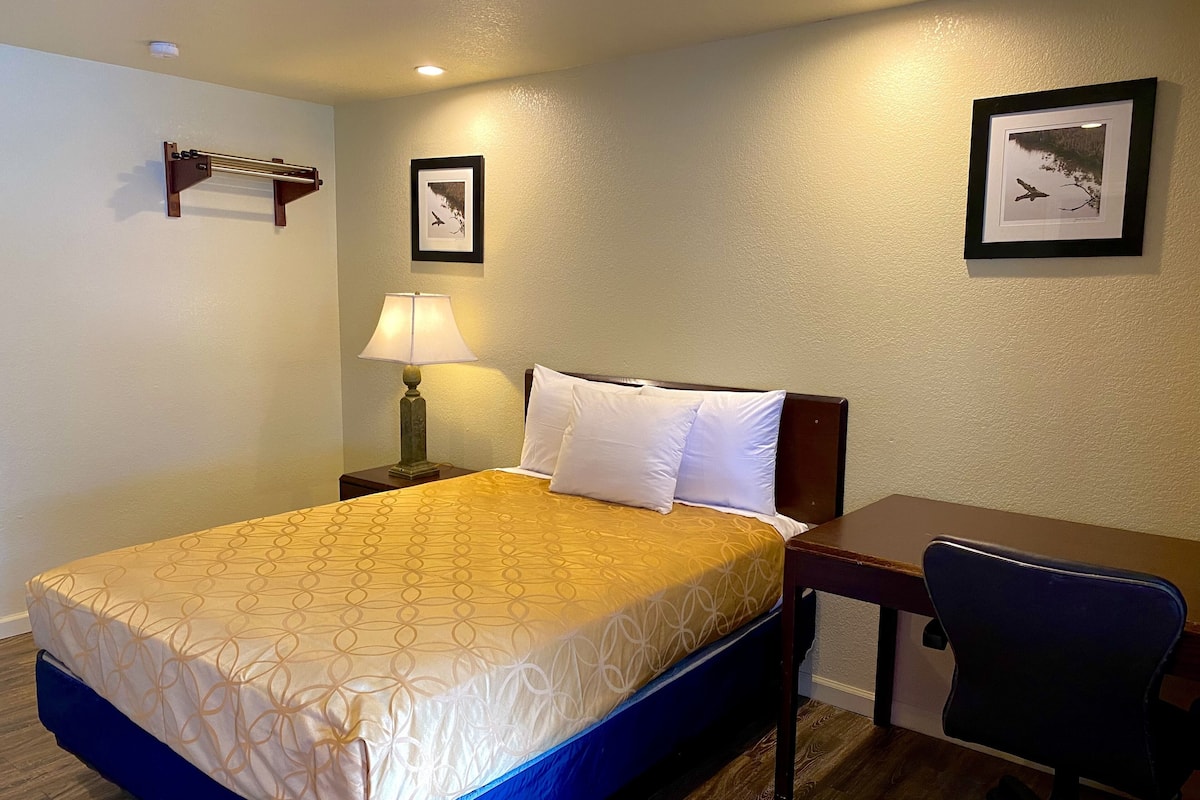 Habitación de hotel con cama, escritorio, silla, lámpara y dos cuadros en la pared.