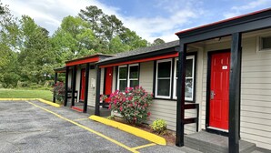A row of motel buildings with red doors and black trim, featuring a parking lot in front. - Whistling Pines - Daily & Extended Stay, Elizabeth City (Elizabeth City)