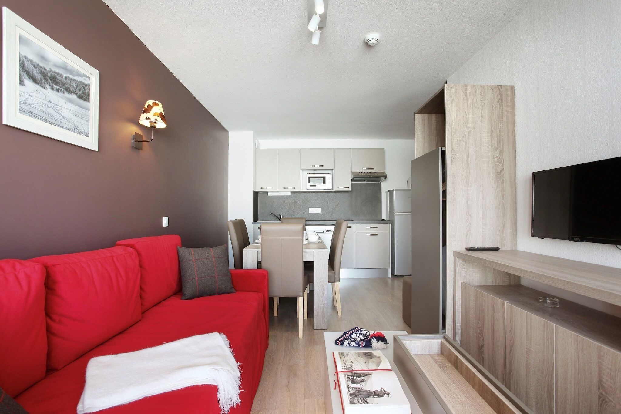 A compact living space with a red sofa, a small kitchen area, and a flat-screen TV mounted on the wall.