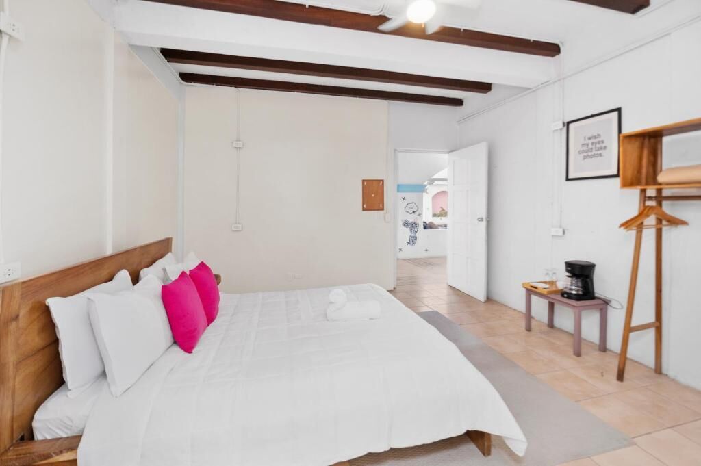 A bedroom with a wooden headboard, white bedding, a ceiling fan, a small table with a coffee maker, and a wooden easel.