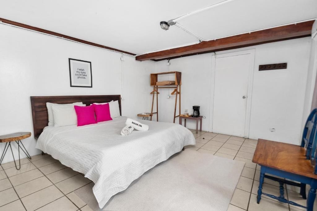 A bedroom with a bed, a chair, a small table, a mirror, and a door.