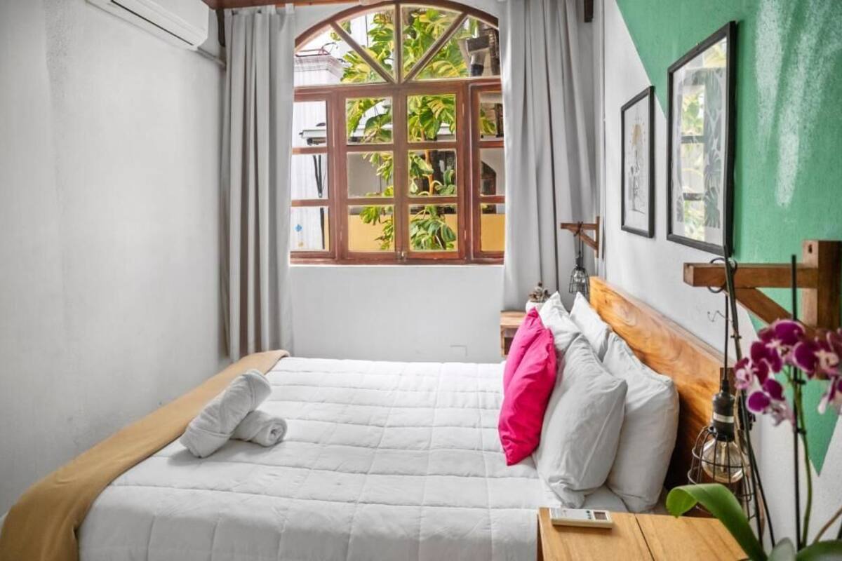 Una cama bien hecha con ropa de cama blanca y un cojín rosa, cabecera de madera, mesita de noche con lámpara y una ventana con cortinas.