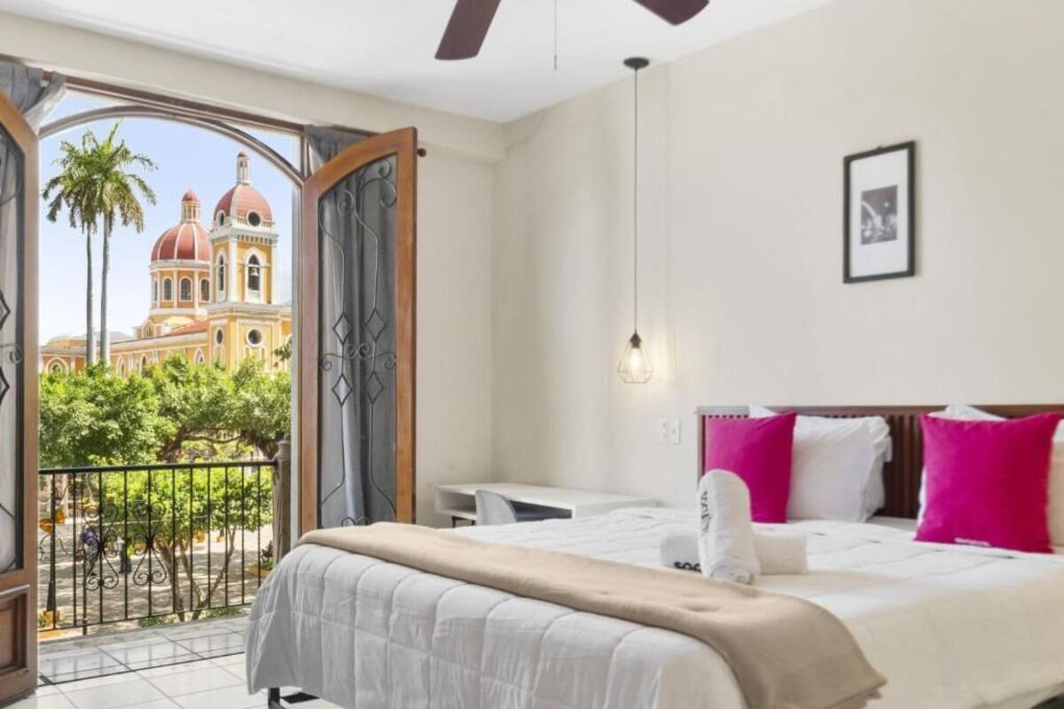A hotel room with a bed, a desk, a chair, a balcony with a view of a building, and a framed picture on the wall.