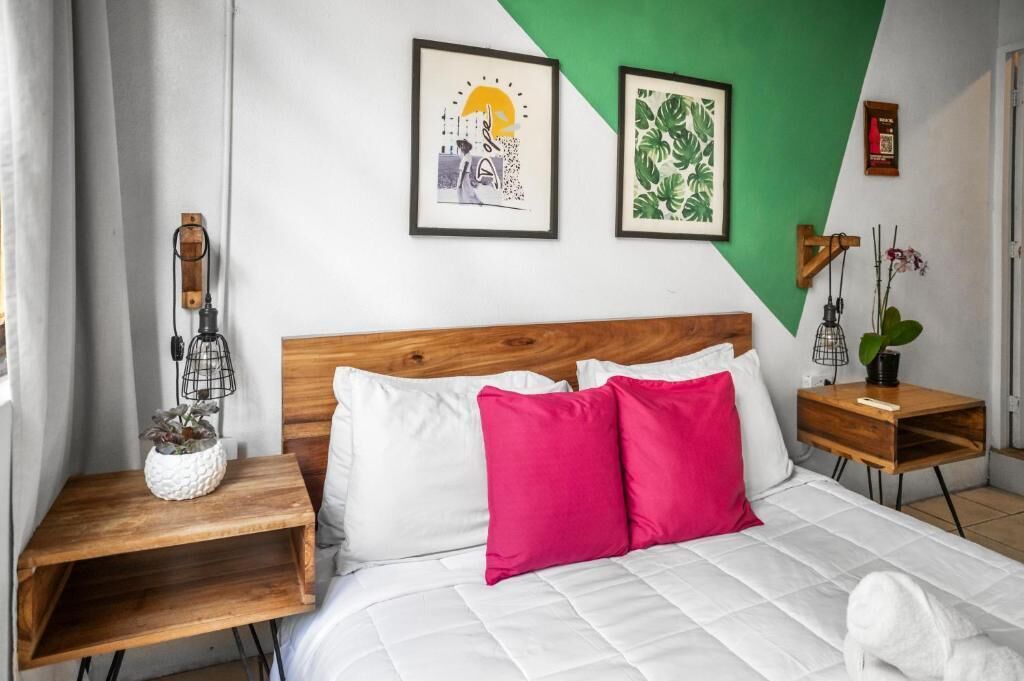 A hotel room with a wooden headboard, white bedding, a red pillow, a small wooden nightstand, a framed picture, and a green accent wall.
