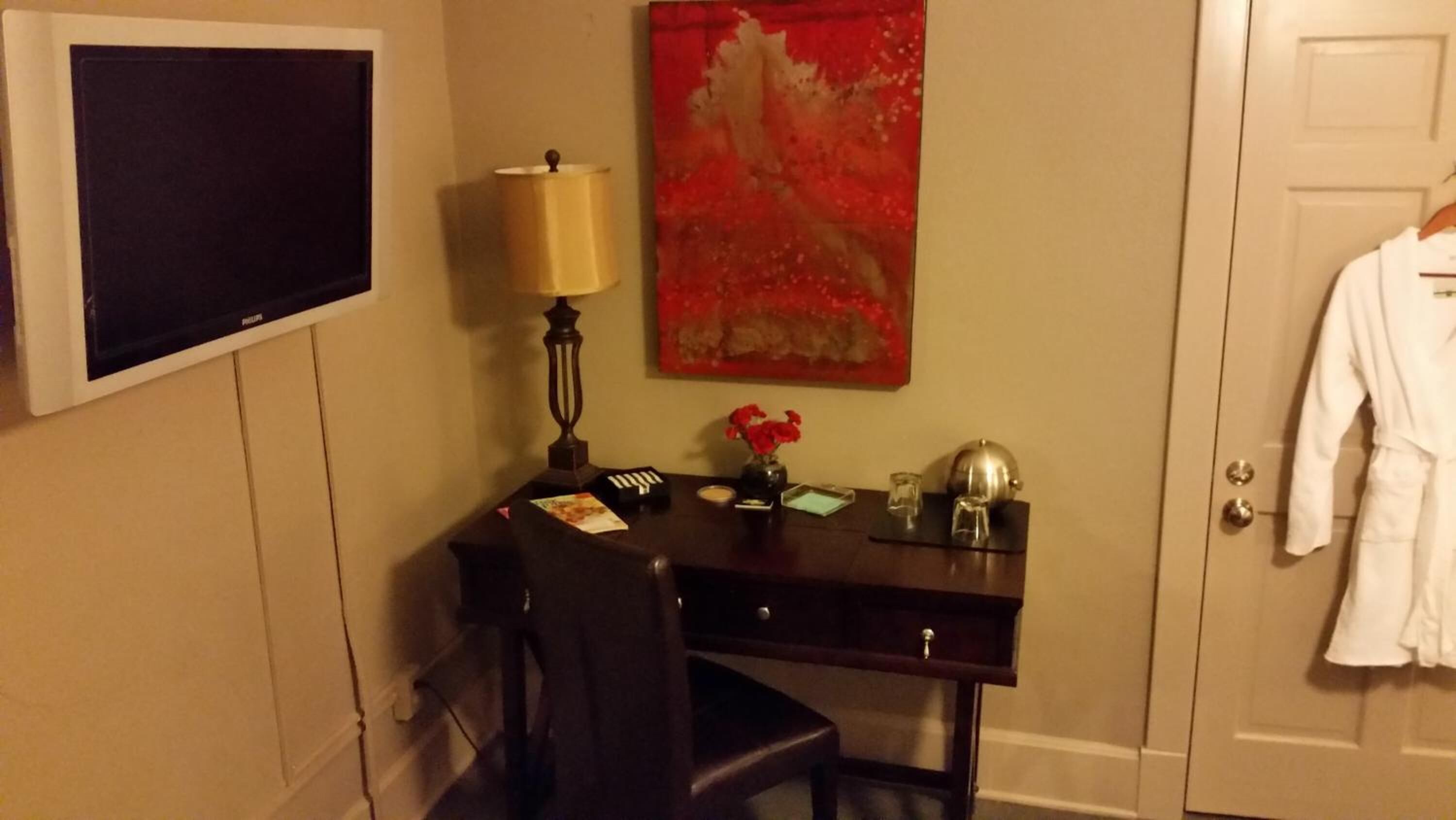 Une chambre d’hôtel équipée d’un bureau, d’une lampe, d’un tableau, d’un vase avec des fleurs, d’une robe suspendue et d’un téléviseur.