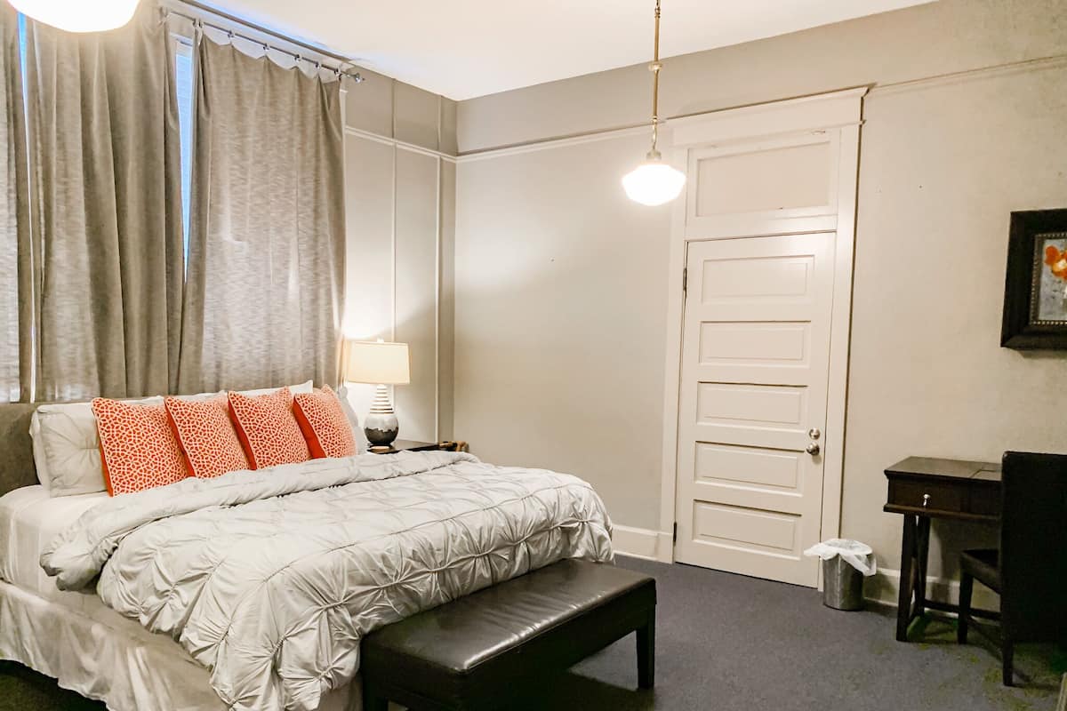 A bedroom with a bed, bench, desk, chair, lamp, and curtains.