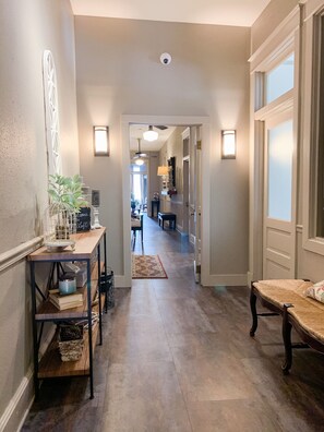 A hallway with a wooden bench, a table with decorative items, and a rug leading to a room with a piano. - The Loft on the Square Guest Suites (Magnolia)