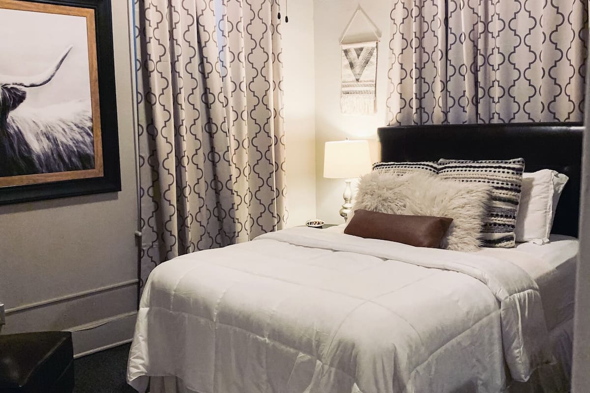 A neatly made bed with a white comforter and pillows, a patterned rug, and a framed picture on the wall.