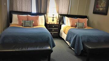 A double room with two beds, each with a blue bedspread and patterned pillows. There are two nightstands with lamps, a small table between the beds, and a bench at the foot of each bed.