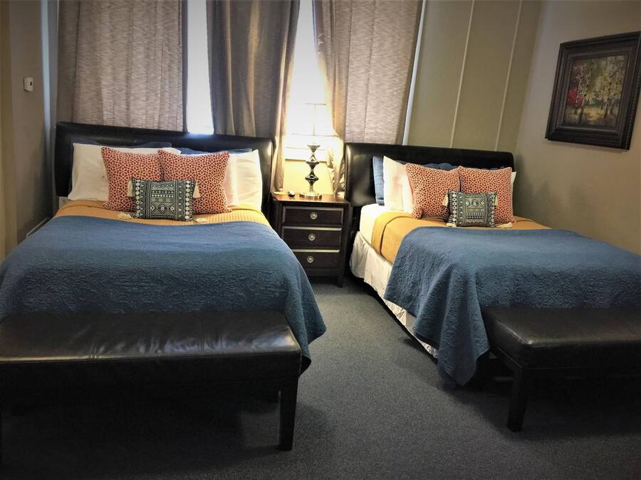 A double room with two beds, each with a blue bedspread and patterned pillows. There are two nightstands with lamps, a small table between the beds, and a bench at the foot of each bed.