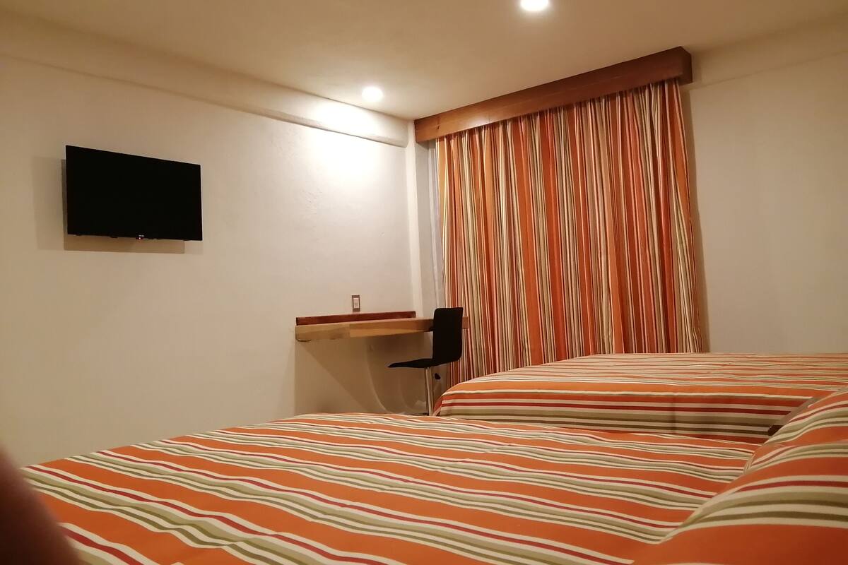 A hotel room with two beds, a flat-screen TV mounted on the wall, a desk with a chair, and a window with striped curtains.
