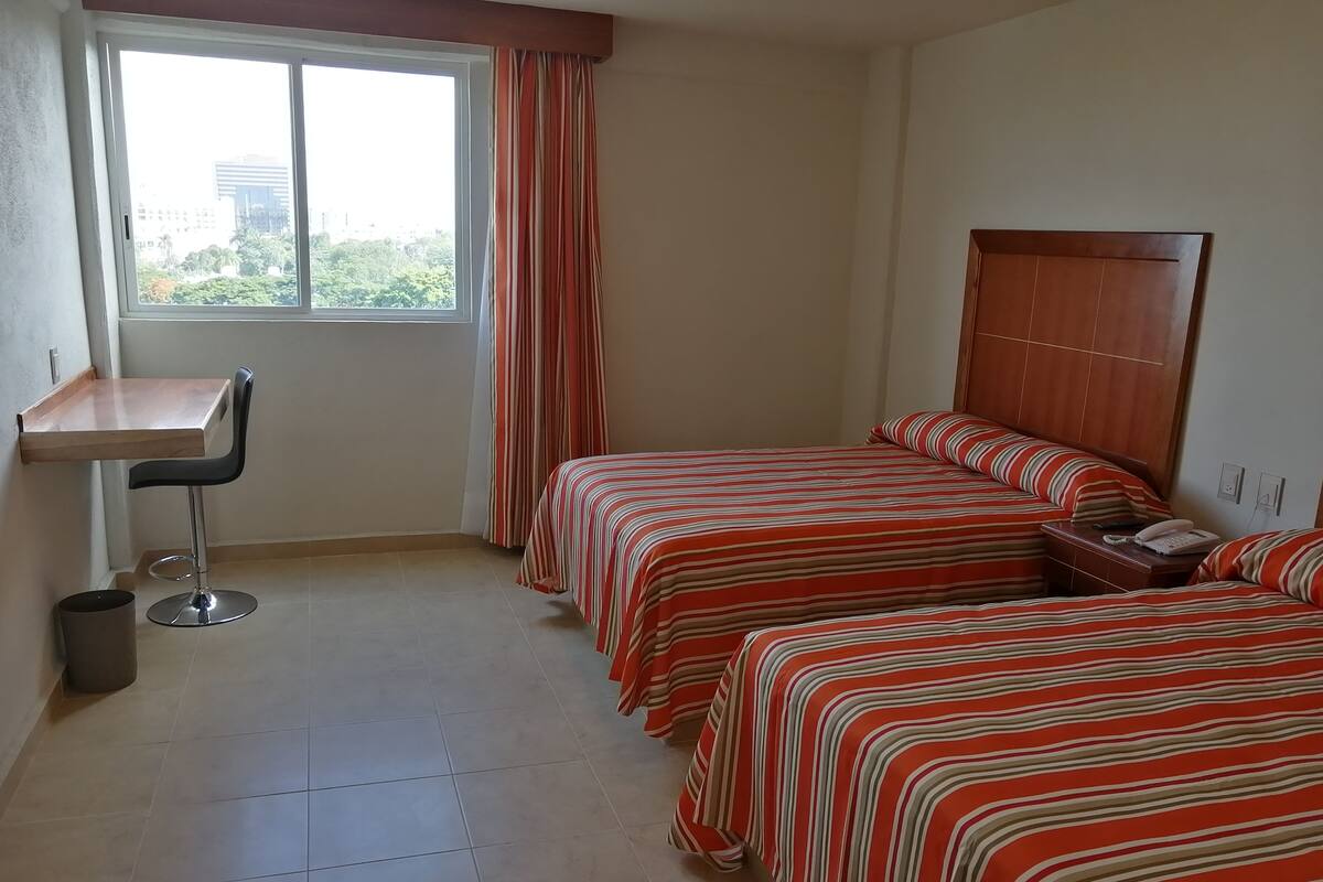 A hotel room with two beds, a small desk, and a window with curtains.