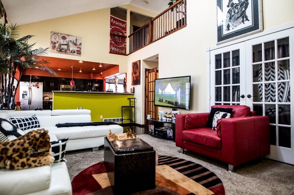 A spacious living area with a white sofa, red armchair, and a TV mounted on the wall. A kitchen is visible in the background with a green countertop.