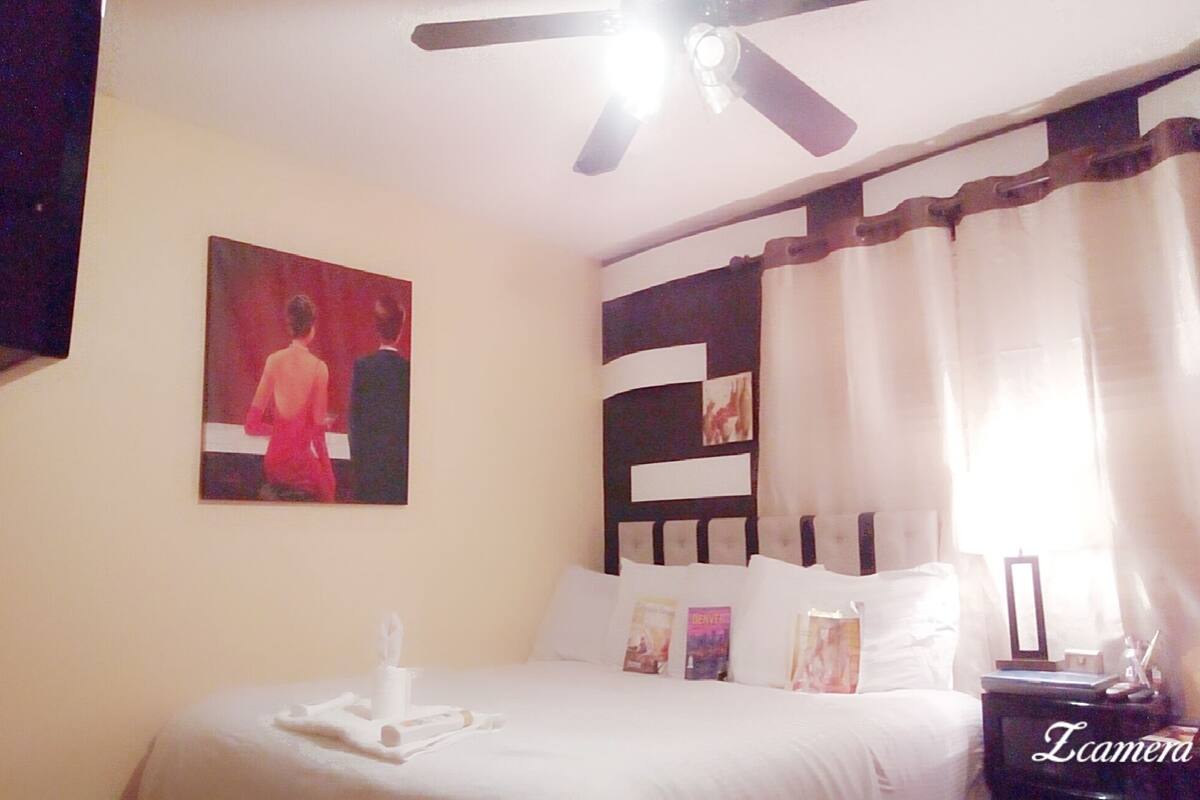 Una habitación de hotel con cama, ventilador de techo, un cuadro en la pared y una ventana con cortinas.