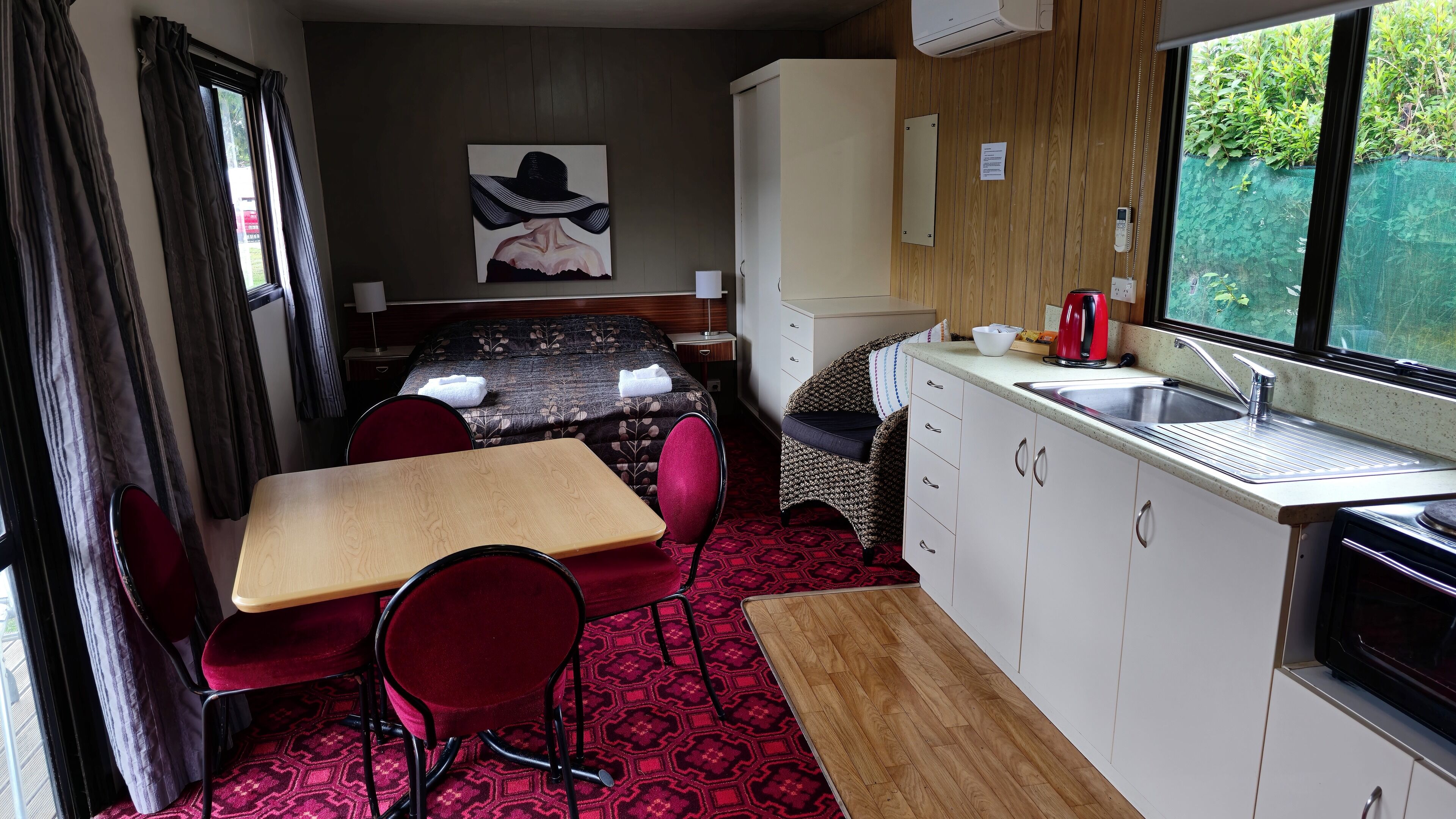 Une pièce compacte comprenant un lit, une petite kitchenette, un coin repas avec une table et des chaises, et une fenêtre donnant sur un espace verdoyant.