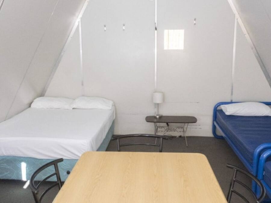 Una habitació petita i triangular amb dos llits, un amb matalàs blau i l'altre amb matalàs blanc. Al centre hi ha una taula de fusta i una petita escriptori amb una làmpada.
