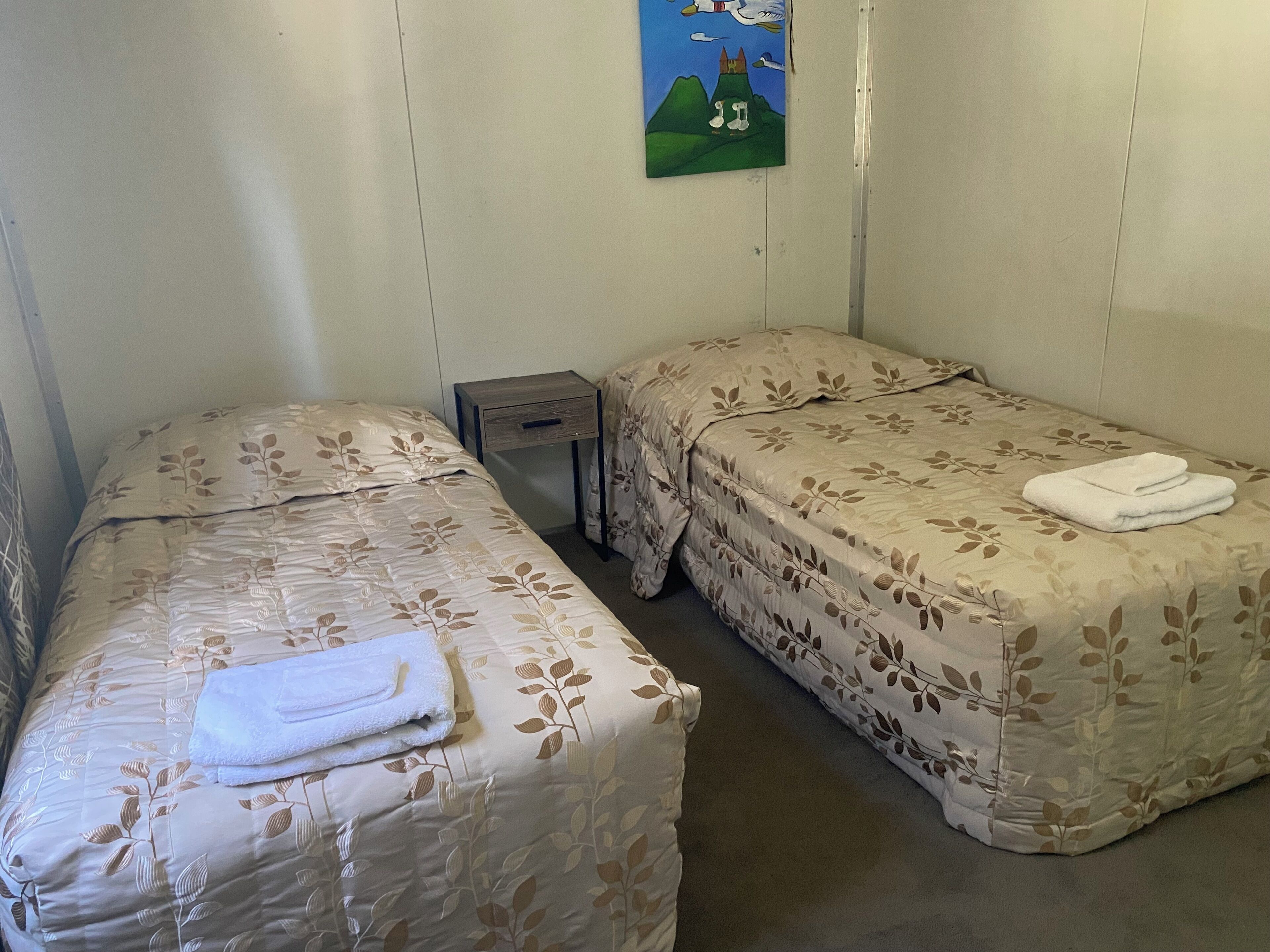 A small, clean room with two single beds, a nightstand, and a painting on the wall.