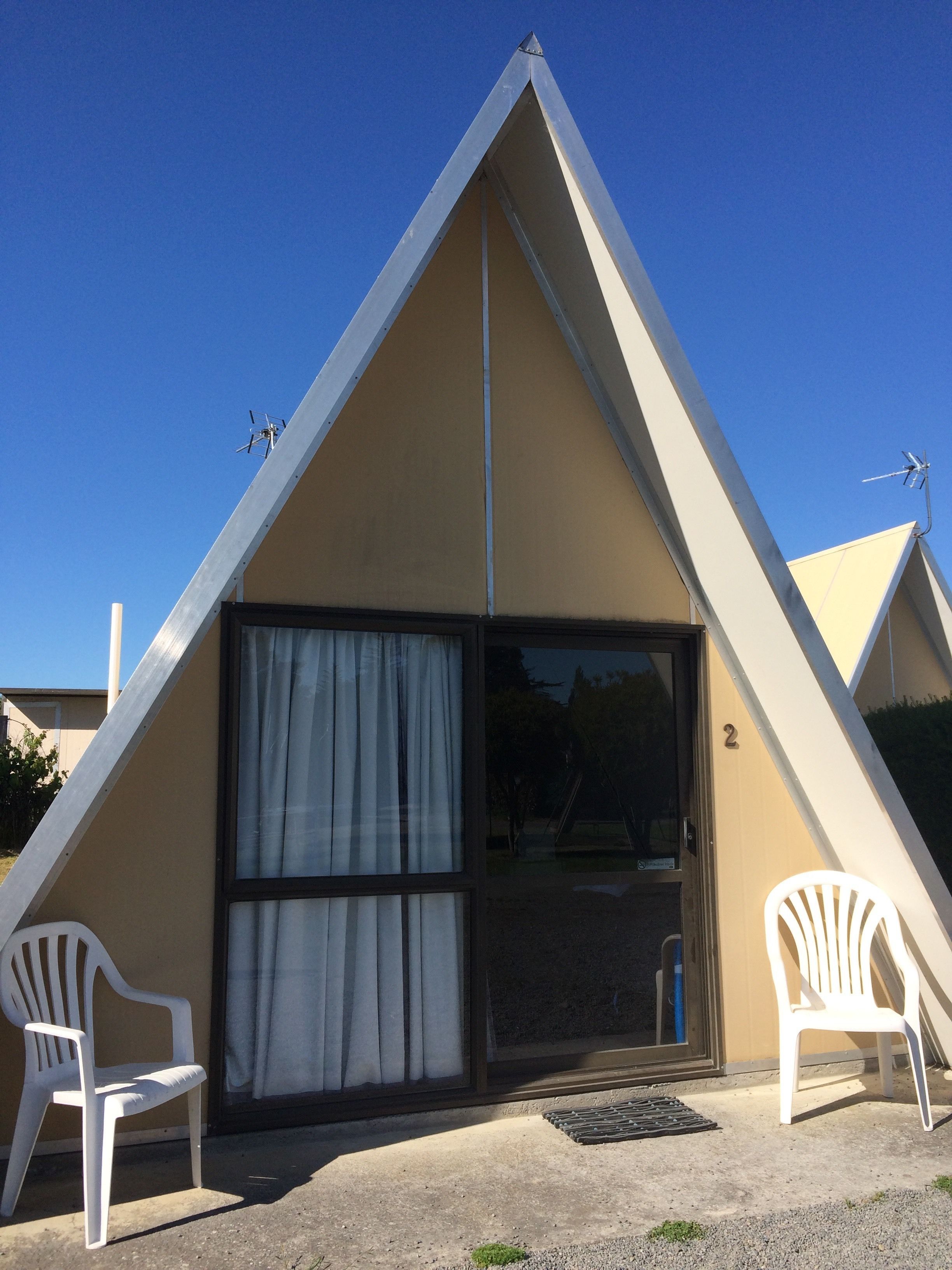 Un edificio triangular con un amplio ventanal corredero, dos sillas de plástico blanco y el número 2 en la puerta.