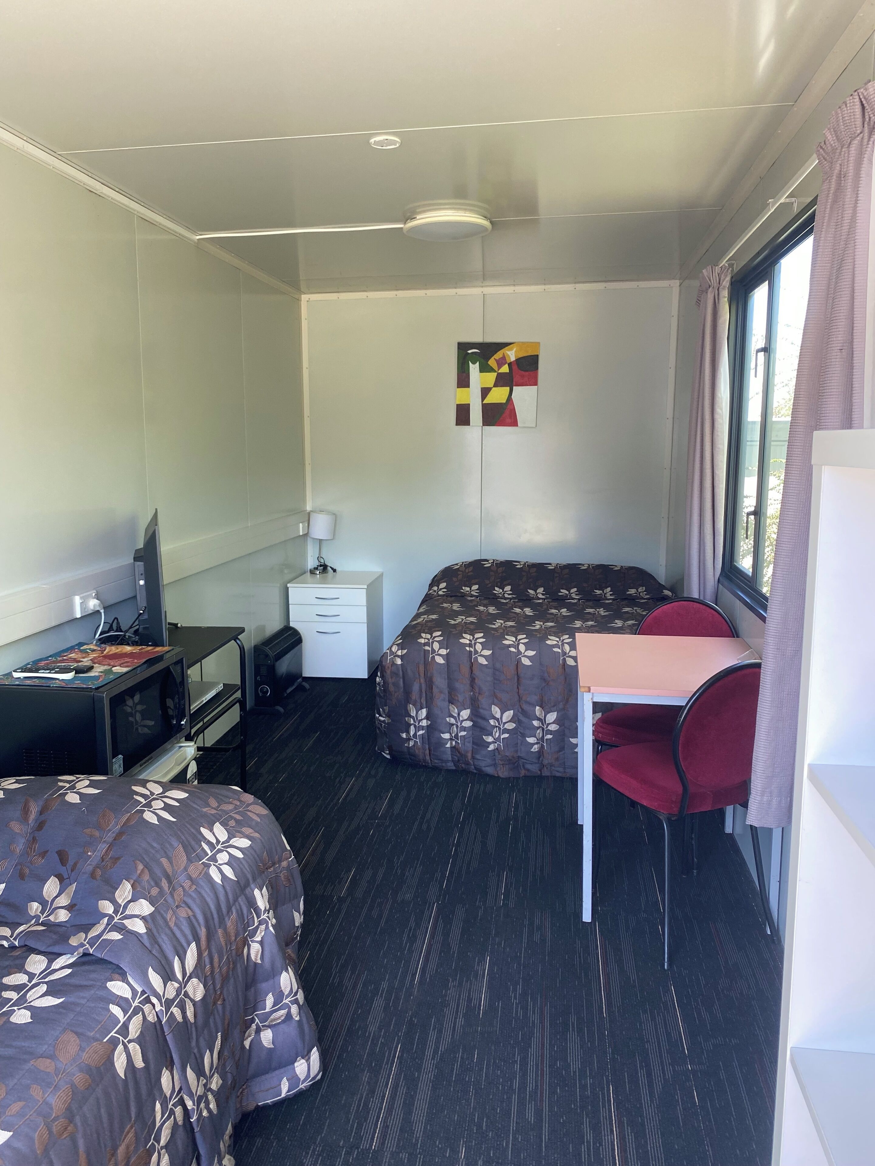 Una habitación pequeña con dos camas, un escritorio, televisor y ventana con cortinas.