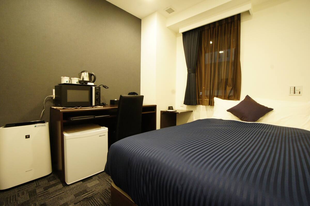 Habitación de hotel con cama, escritorio con microondas, refrigerador y ventana con cortinas.