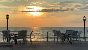 Una terraza frente al mar con sillas y mesas, barandilla y un atardecer sobre el océano.