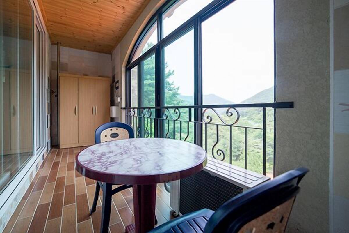 A balcony with a round table and chairs, a wooden ceiling, and a view of greenery and hills.