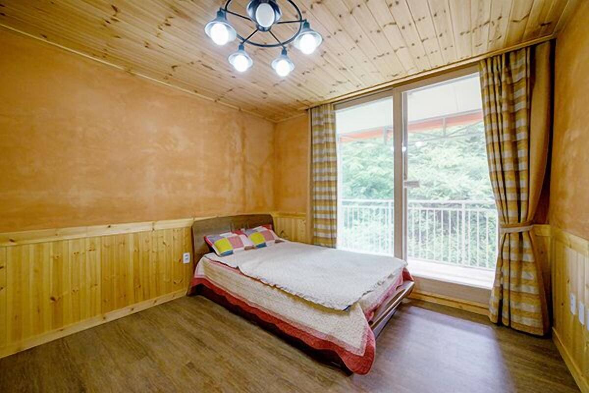 A wooden-paneled bedroom with a bed, a window with curtains, and a view of greenery outside.