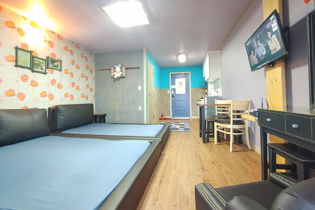 A room with a bed, a desk, and a chair. There is a door with a sign, a wall-mounted TV, and a small table.
