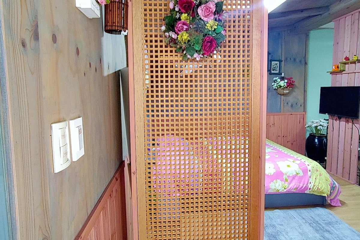 A wooden room with a bed, a wooden partition with a floral arrangement, and a small shelf with flowers.