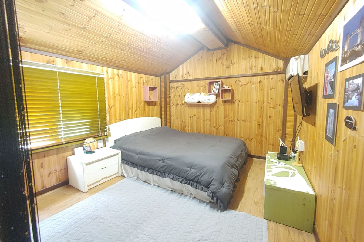 A bedroom with wooden walls, a bed with a grey bedspread, a bedside table, a nightstand, and a green nightstand.